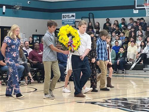 students in gym filled with students and families walking across floor with wreath