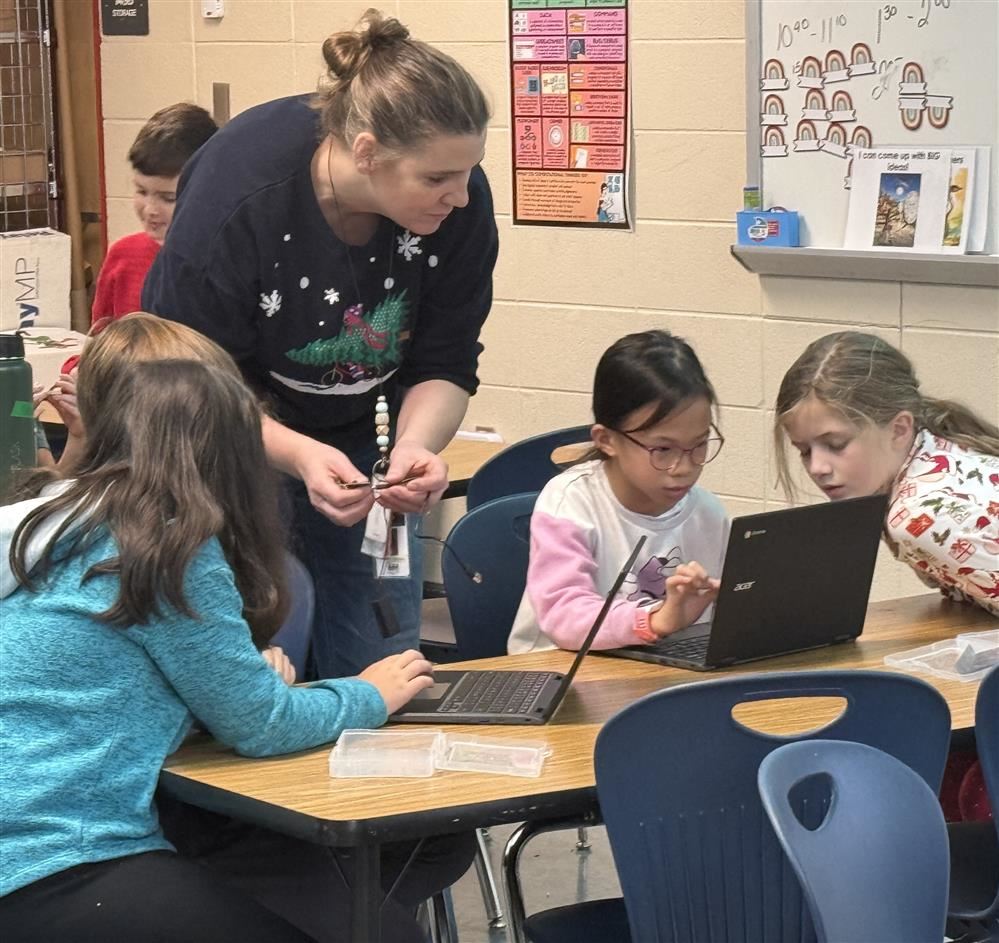  Image of a teacher in front of class addressing students clustered together over a group project
